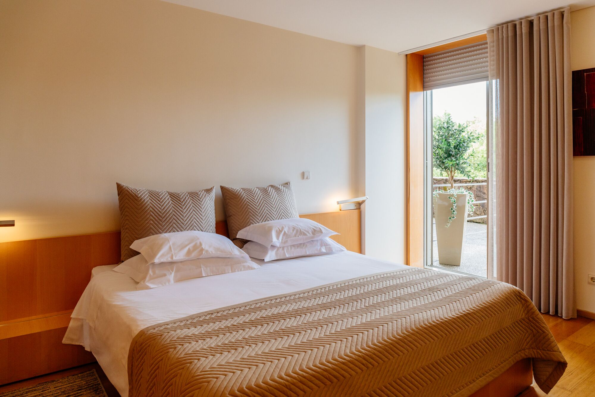 A hotel room with a bed, two pillows, a nightstand, a window with curtains, and a view of greenery outside.