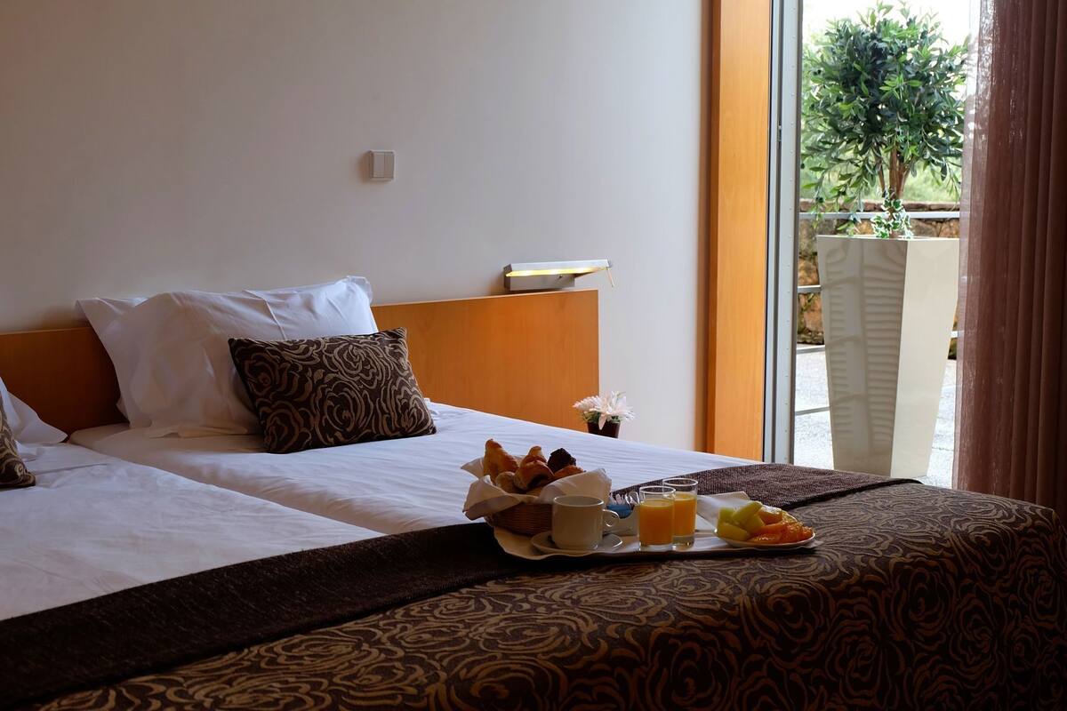 Hotelkamer met twee bedden, een ontbijtkarretje met croissants, sap en fruit, en uitzicht op groen.