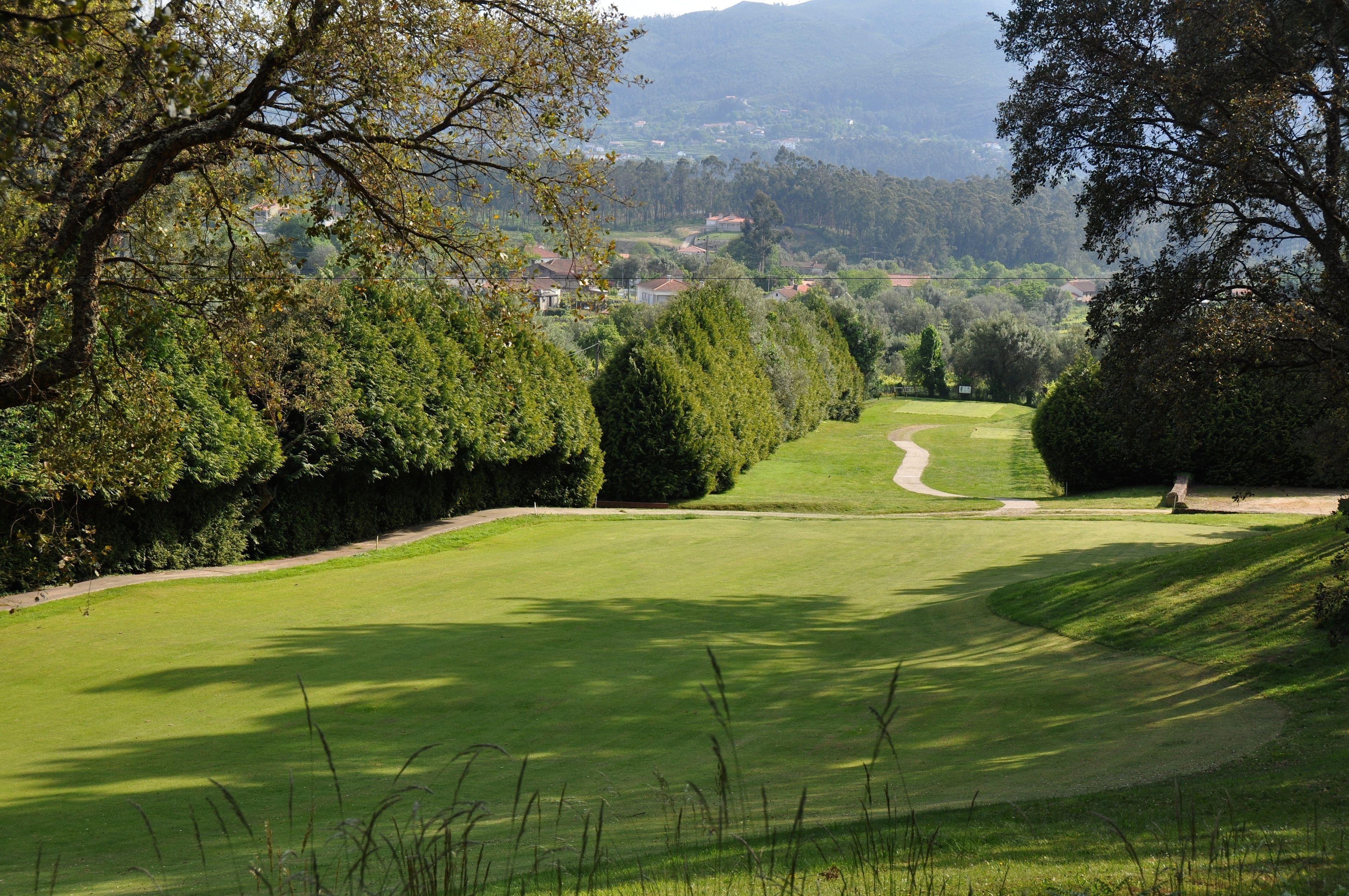 A golf course with a well-maintained fairway, a putting green, and a backdrop of trees and distant houses.