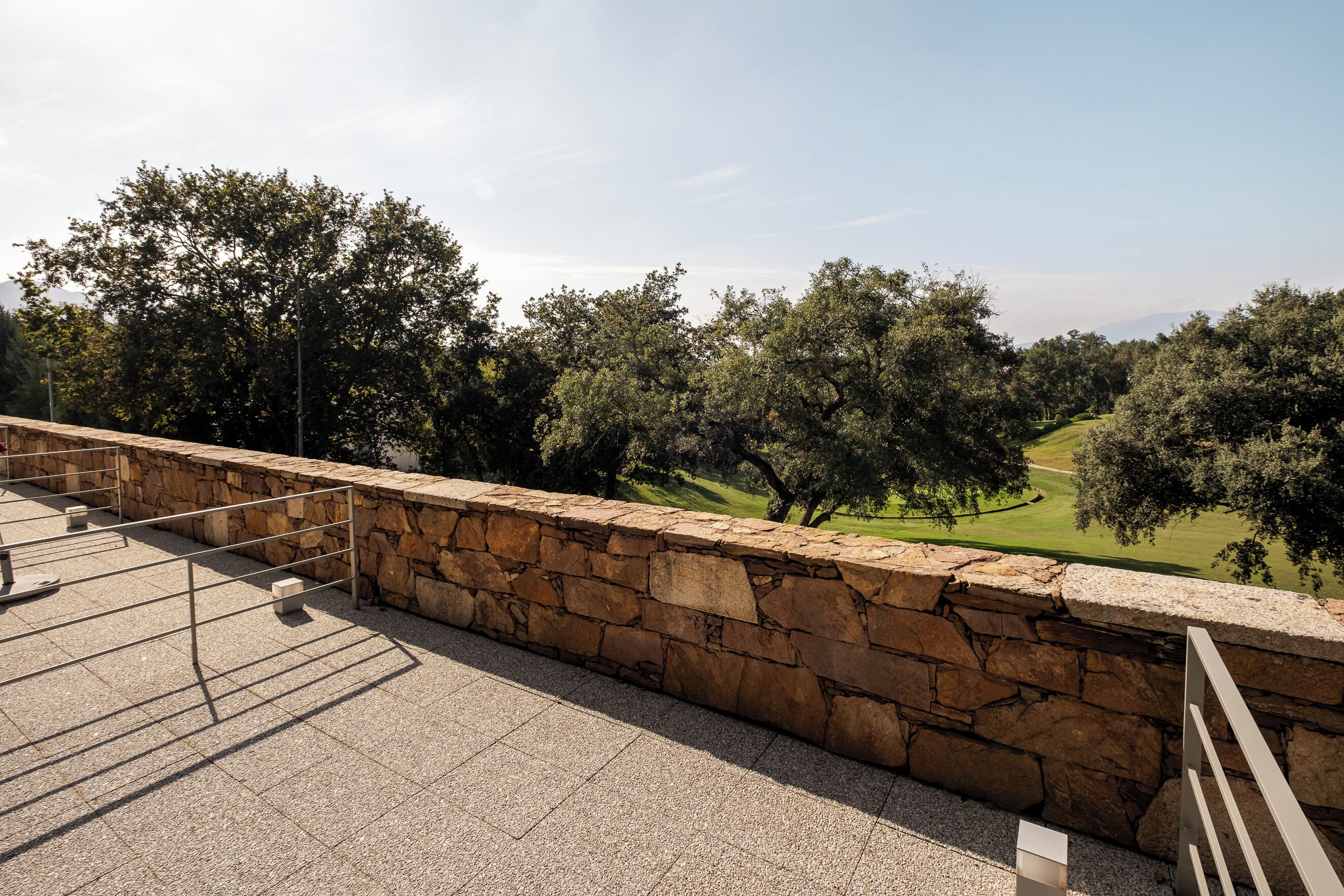 A stone wall with a metal railing overlooking a green landscape with trees.