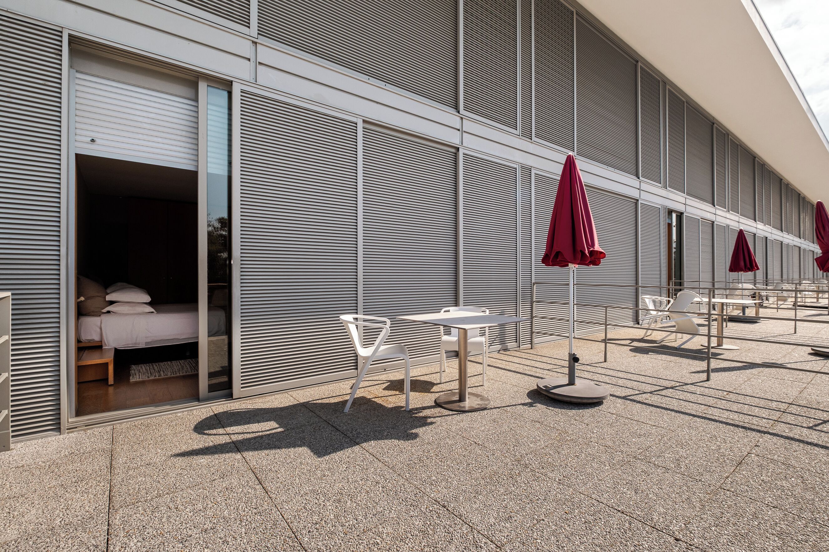 Un edificio moderno con una zona de estar al aire libre techada, con sillas blancas y sombrillas rojas, y se puede ver un dormitorio con cama y almohadas.