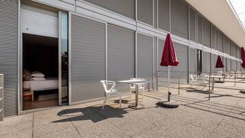 Un edificio moderno con una zona de estar al aire libre techada, con sillas blancas y sombrillas rojas, y se puede ver un dormitorio con cama y almohadas.