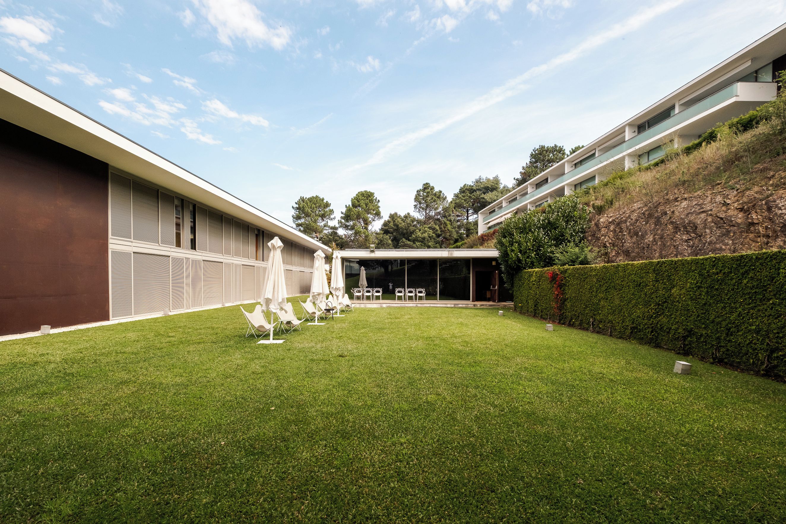 A modern building with a flat roof, a covered outdoor seating area with chairs and umbrellas, and a well-maintained lawn.