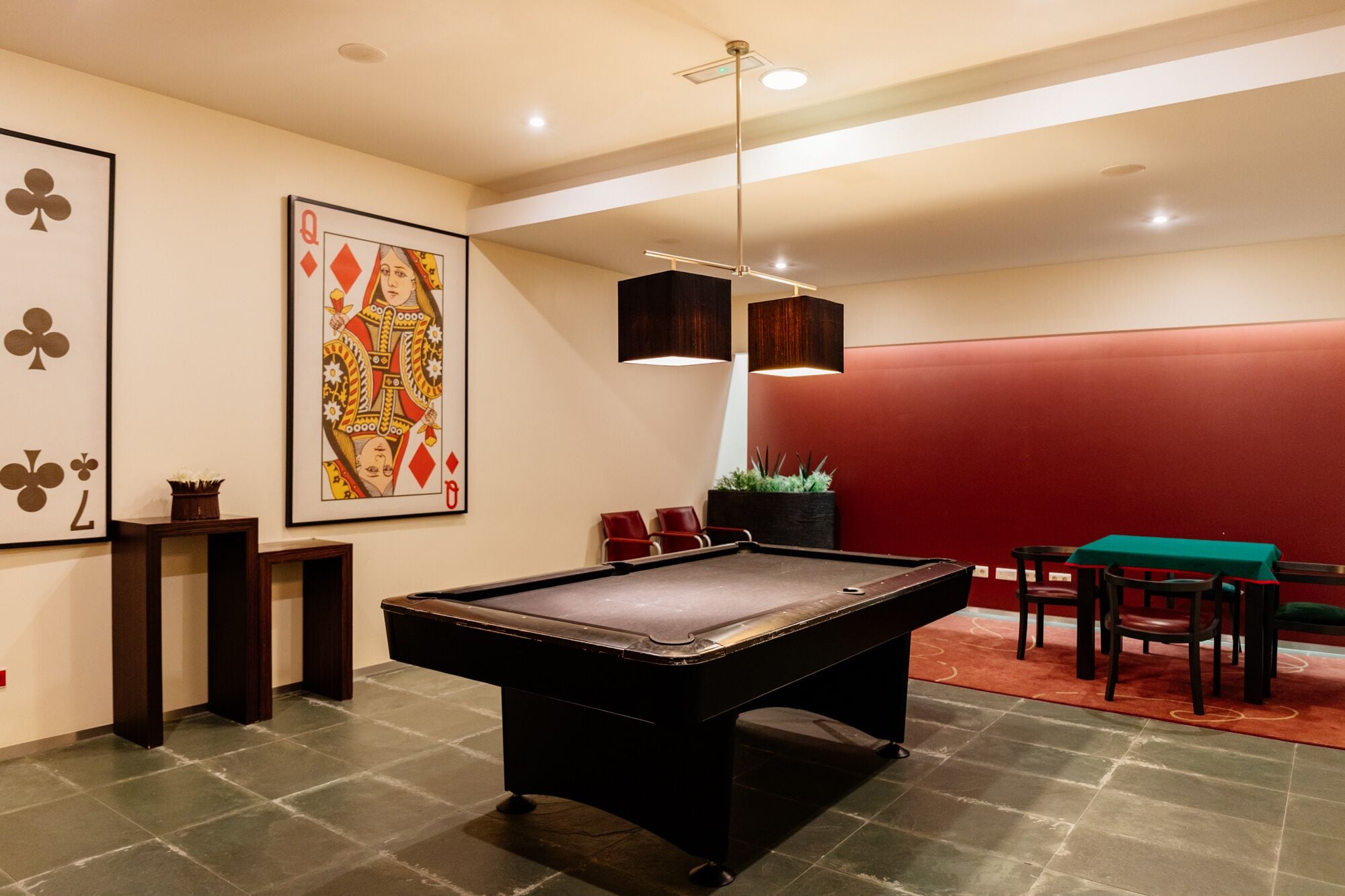 A billiards table in a room with a red accent wall, a large framed playing card, and a potted plant.