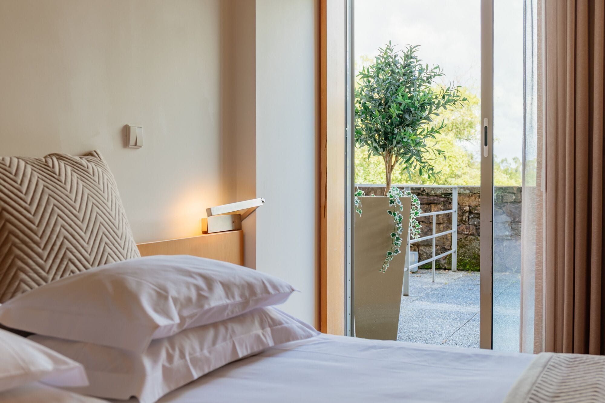Habitación de hotel con cama, mesita de noche, lámpara de pared y vistas a un balcón con vegetación.