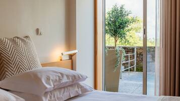 Habitación de hotel con cama, mesita de noche, lámpara de pared y vistas a un balcón con vegetación.