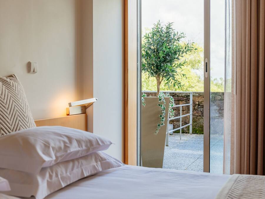 Habitación de hotel con cama, mesita de noche, lámpara de pared y vistas a un balcón con vegetación.