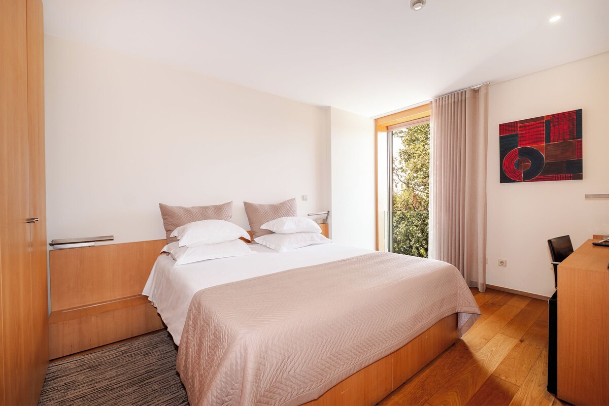 A modern hotel room with a large bed, a desk, and a view of greenery through the window.
