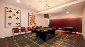 Ein Billardtisch in einem Raum mit roter Wand, einer grünen Spieltischplatte und einer Sitzecke mit Stühlen.