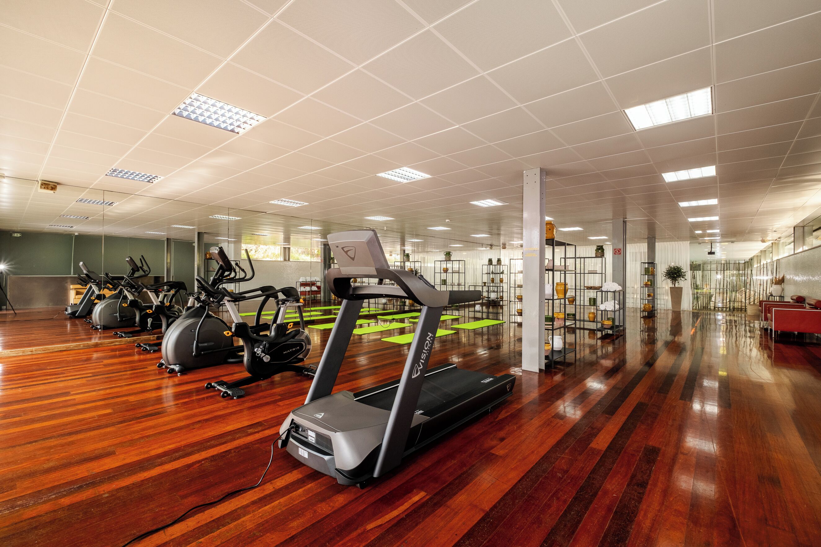 Uma academia ampla com várias esteiras e bicicletas ergométricas, piso de madeira envernizada e teto iluminado com luminárias quadradas.