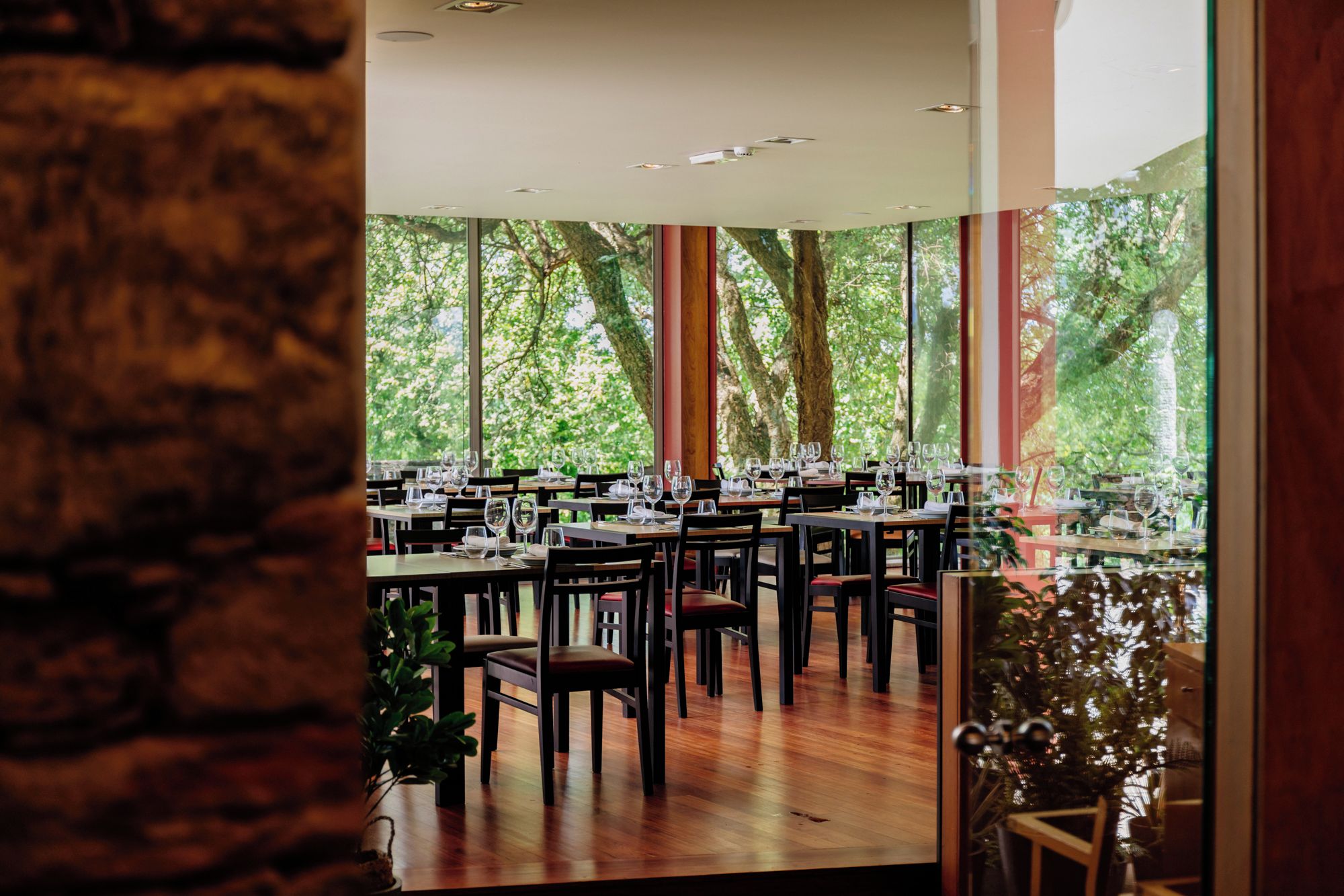 A restaurant with multiple tables and chairs, large windows with a view of trees, and a brick wall.
