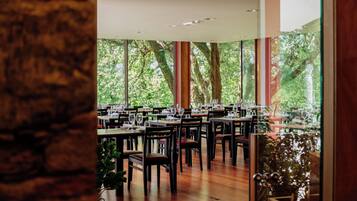 A restaurant with multiple tables and chairs, large windows with a view of trees, and a brick wall.