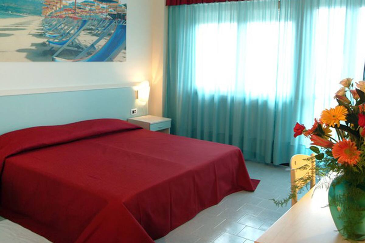 Una camera d'albergo con un letto, un comodino, una lampada, una finestra con tende e un quadro appeso al muro.