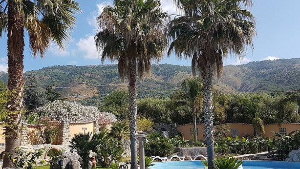 A pool area with palm trees, stone architecture, and a mountainous backdrop. - Cliffs Hotel (Joppolo)