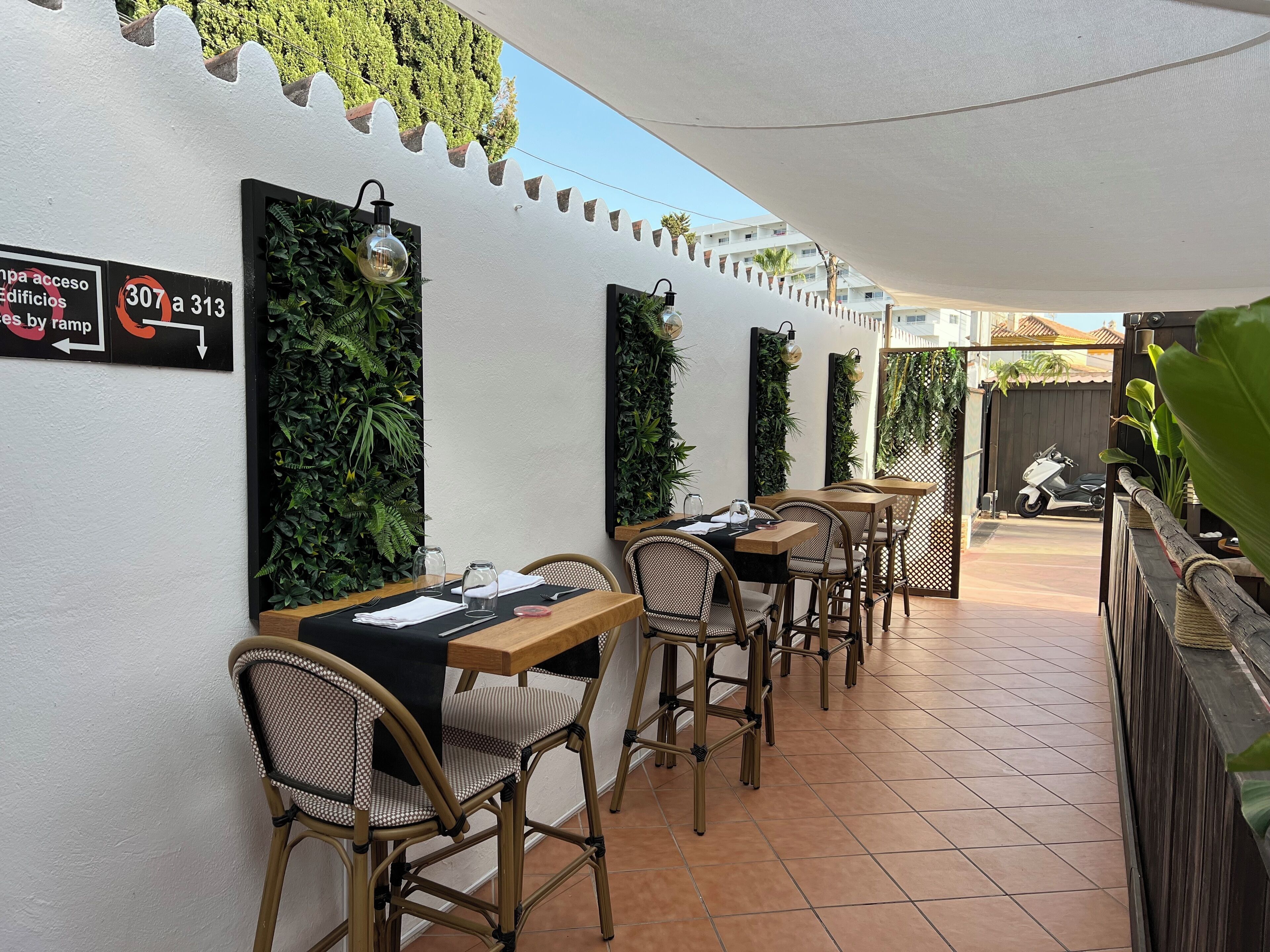 Un patio con mobiliario exterior, mesas y sillas, y un cartel que indica acceso a edificios mediante rampa.