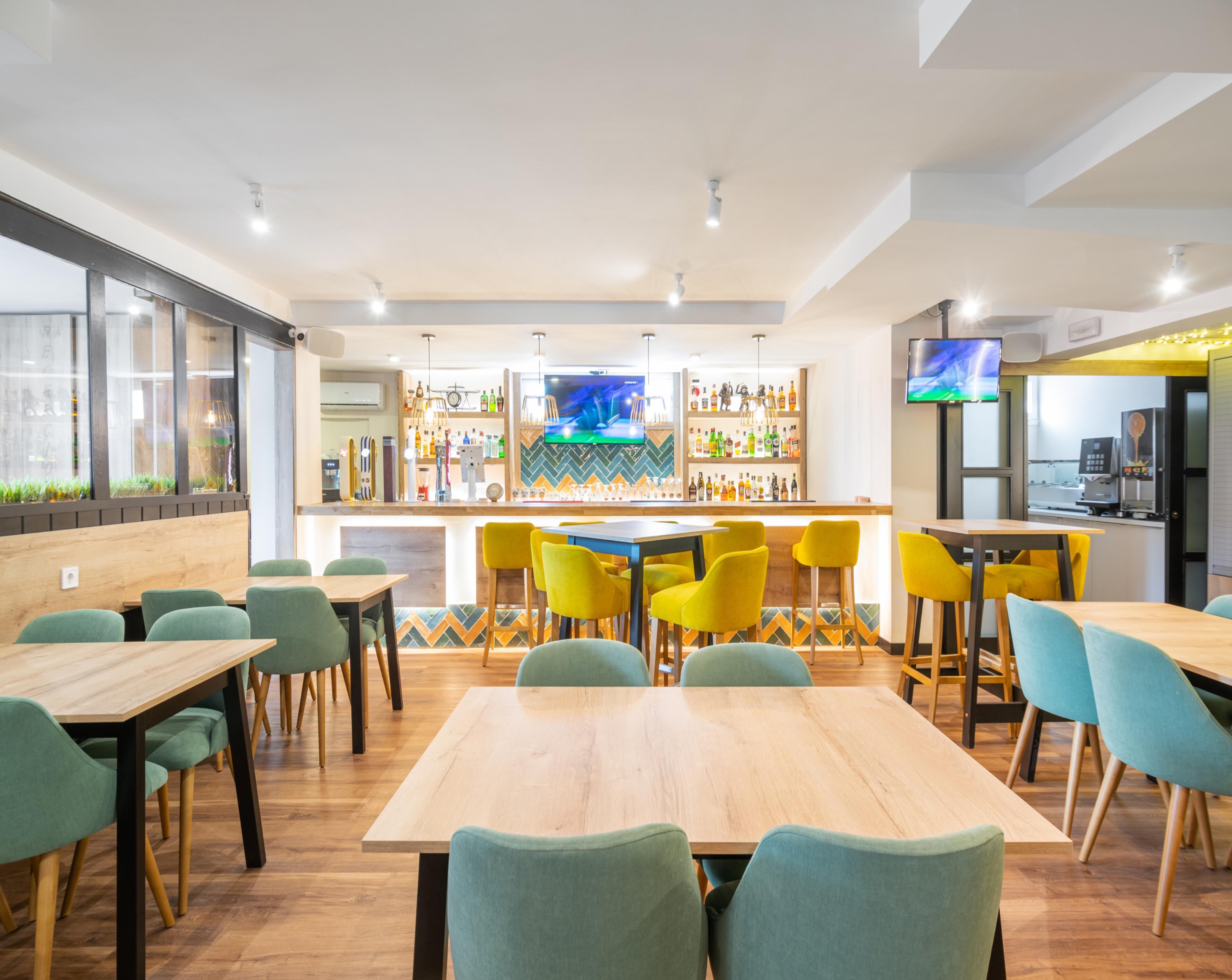 A modern bar area with a counter, bar stools, and a television screen. There are tables and chairs arranged around the bar.
