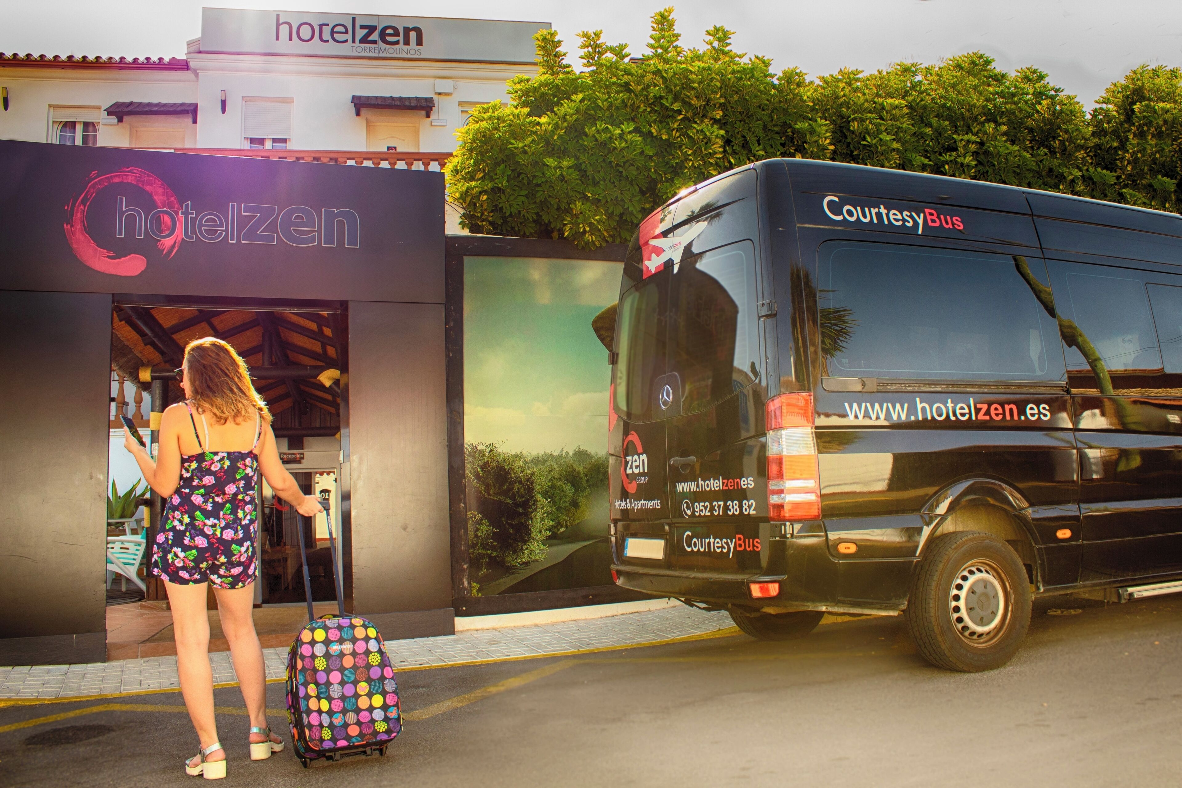 Hoteleingang mit einem Bus, der mit 'Courtesy Bus' beschriftet ist, und einer Person mit einem bunten Koffer.