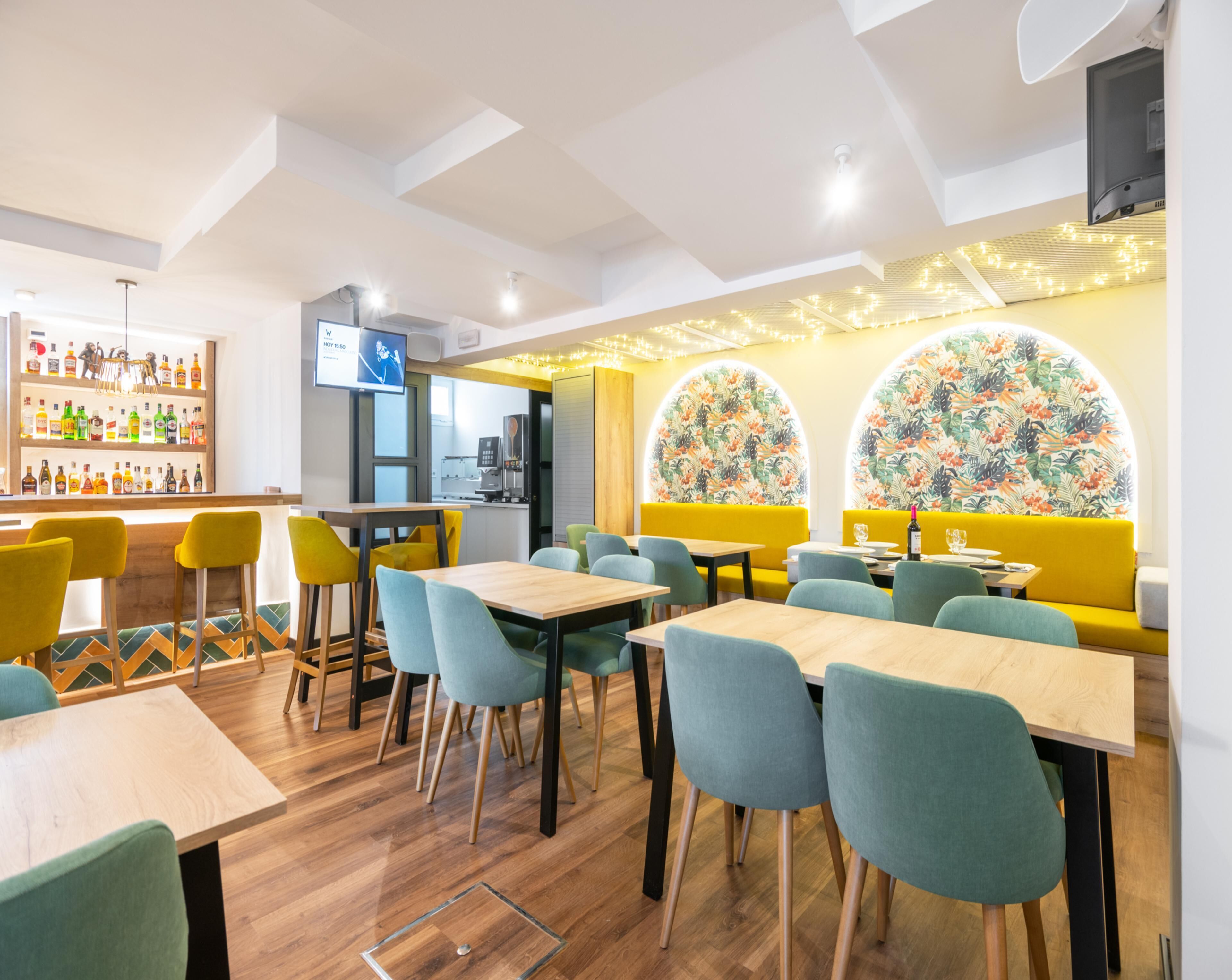 A restaurant with yellow booths, teal chairs, and wooden tables. There is a bar area with a variety of bottles and a TV screen.
