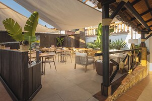 Een terras buiten met houten tafels en stoelen, een rieten dak en tropische planten.