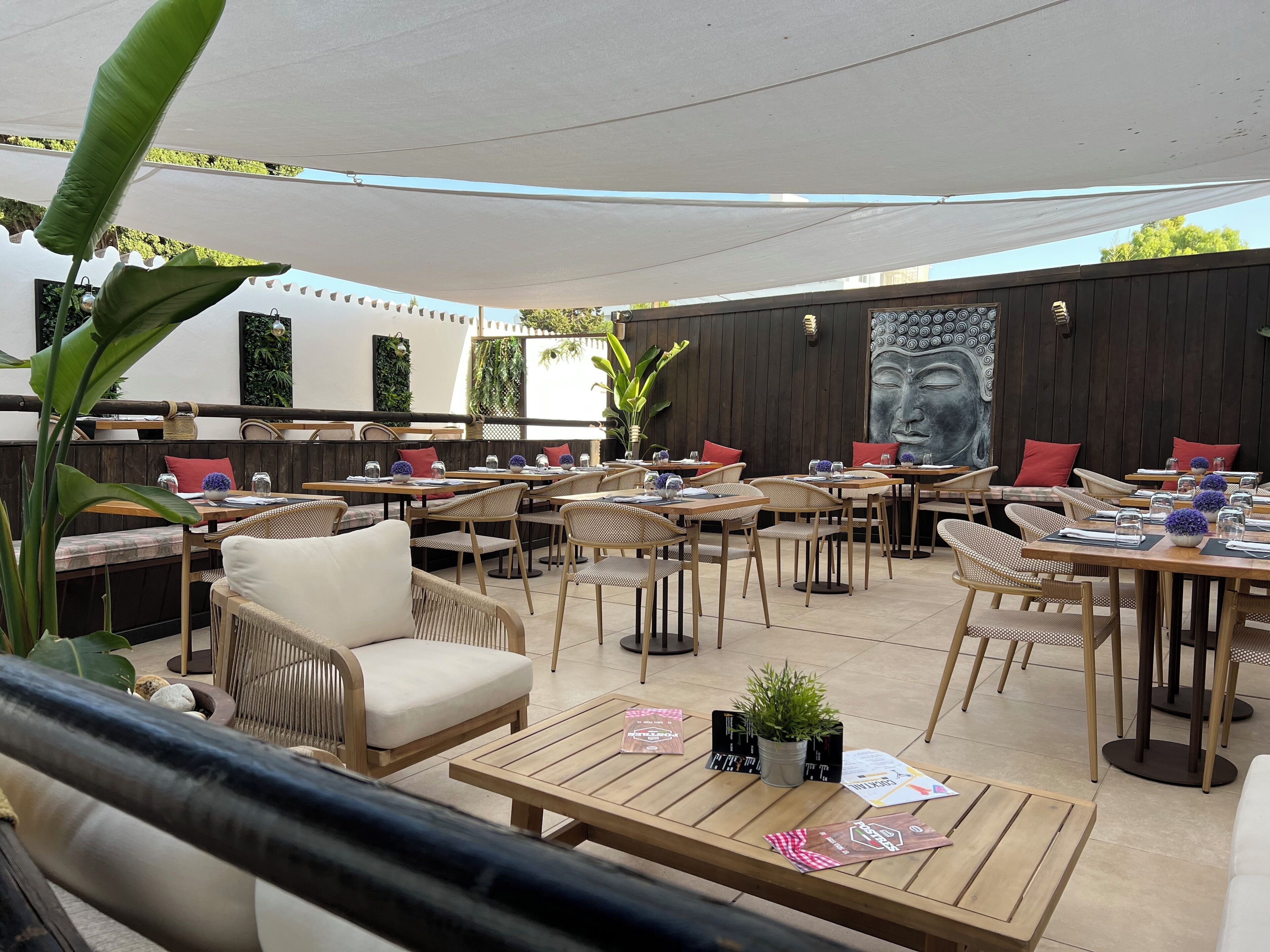 An outdoor dining area with wooden tables and chairs, potted plants, and a Buddha mural on the wall.
