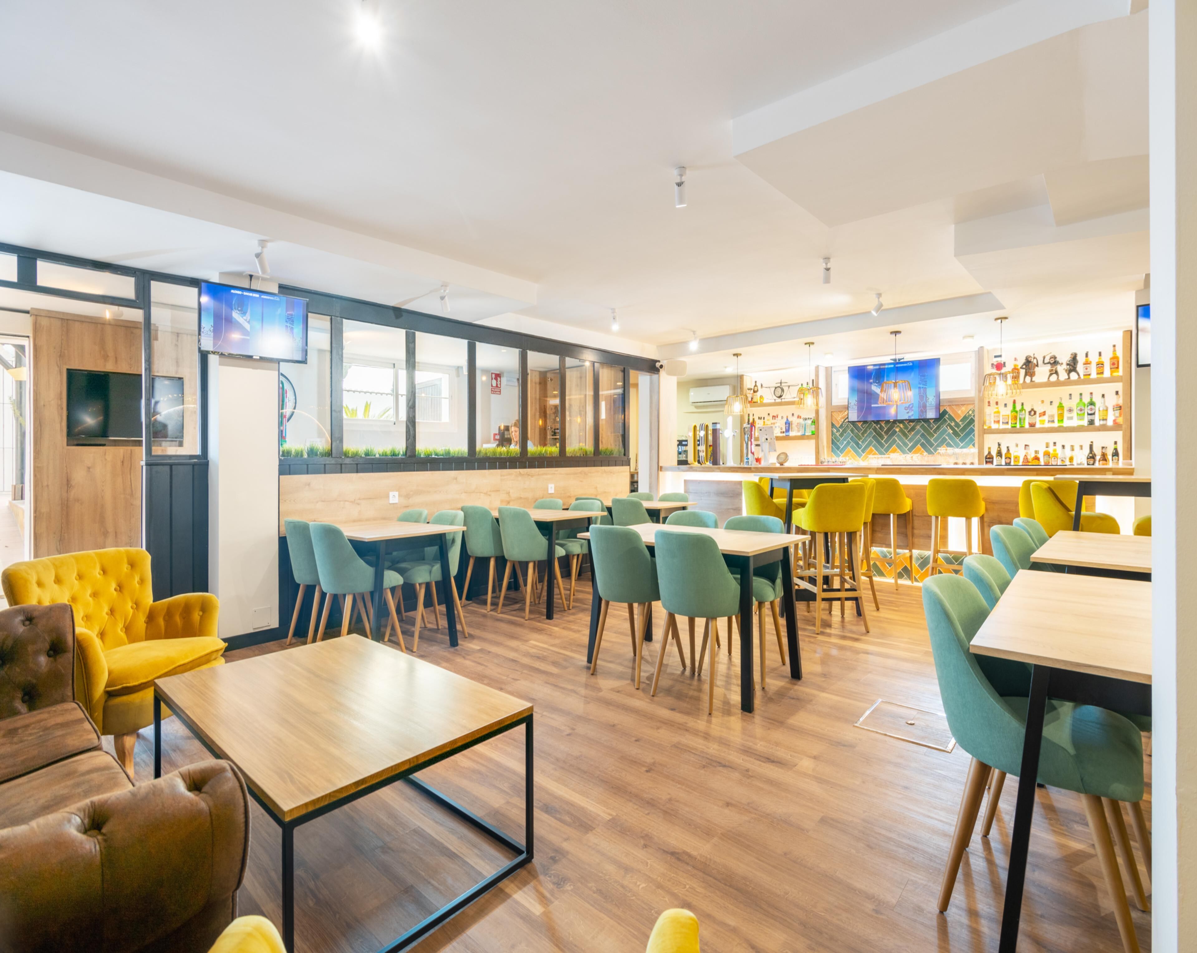 A modern restaurant interior with wooden flooring, green and yellow chairs, and a bar area with shelves of bottles.