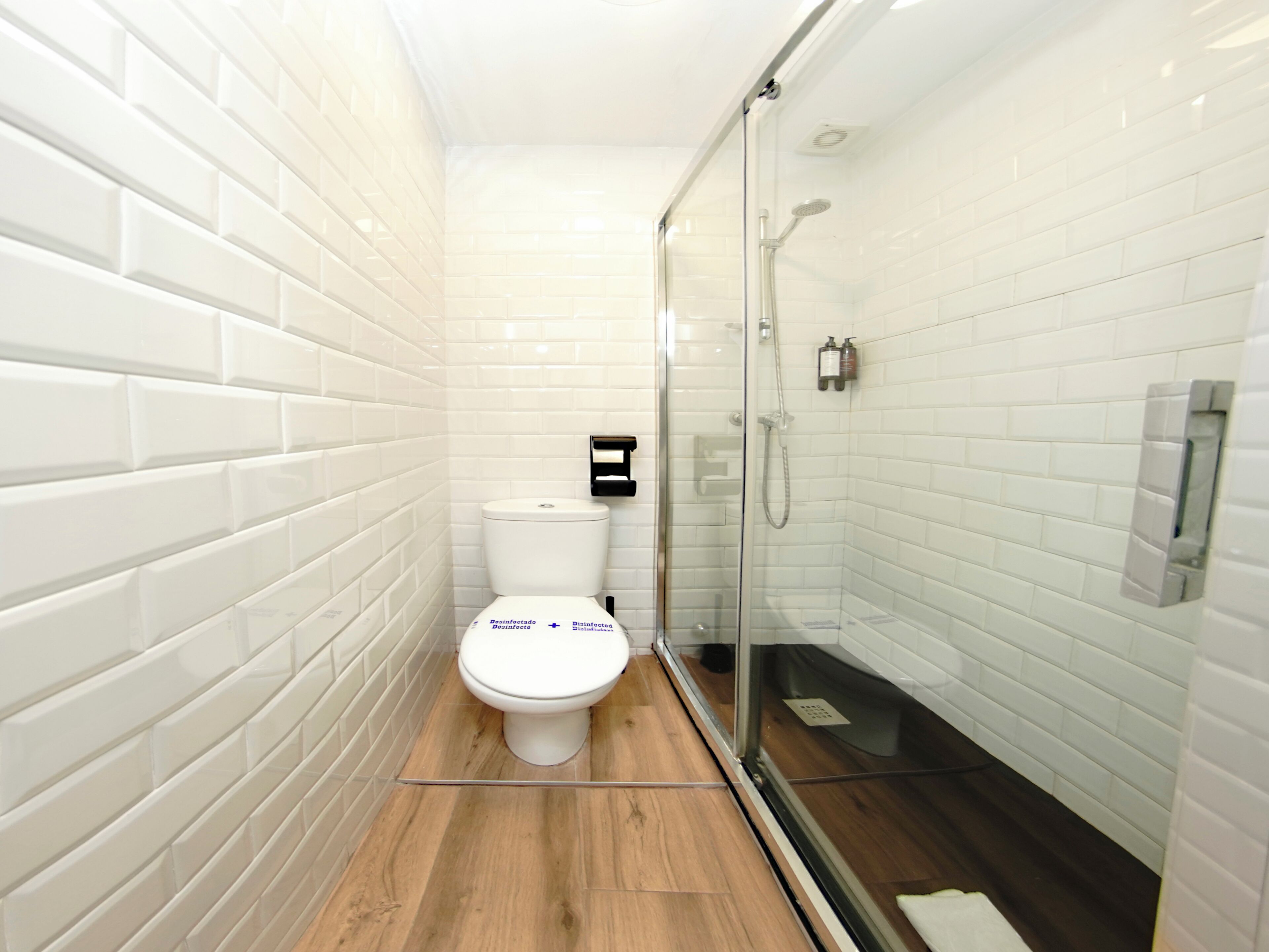 Un baño moderno con azulejos blancos tipo metro, un inodoro blanco, ducha envuelta en vidrio y piso de madera.