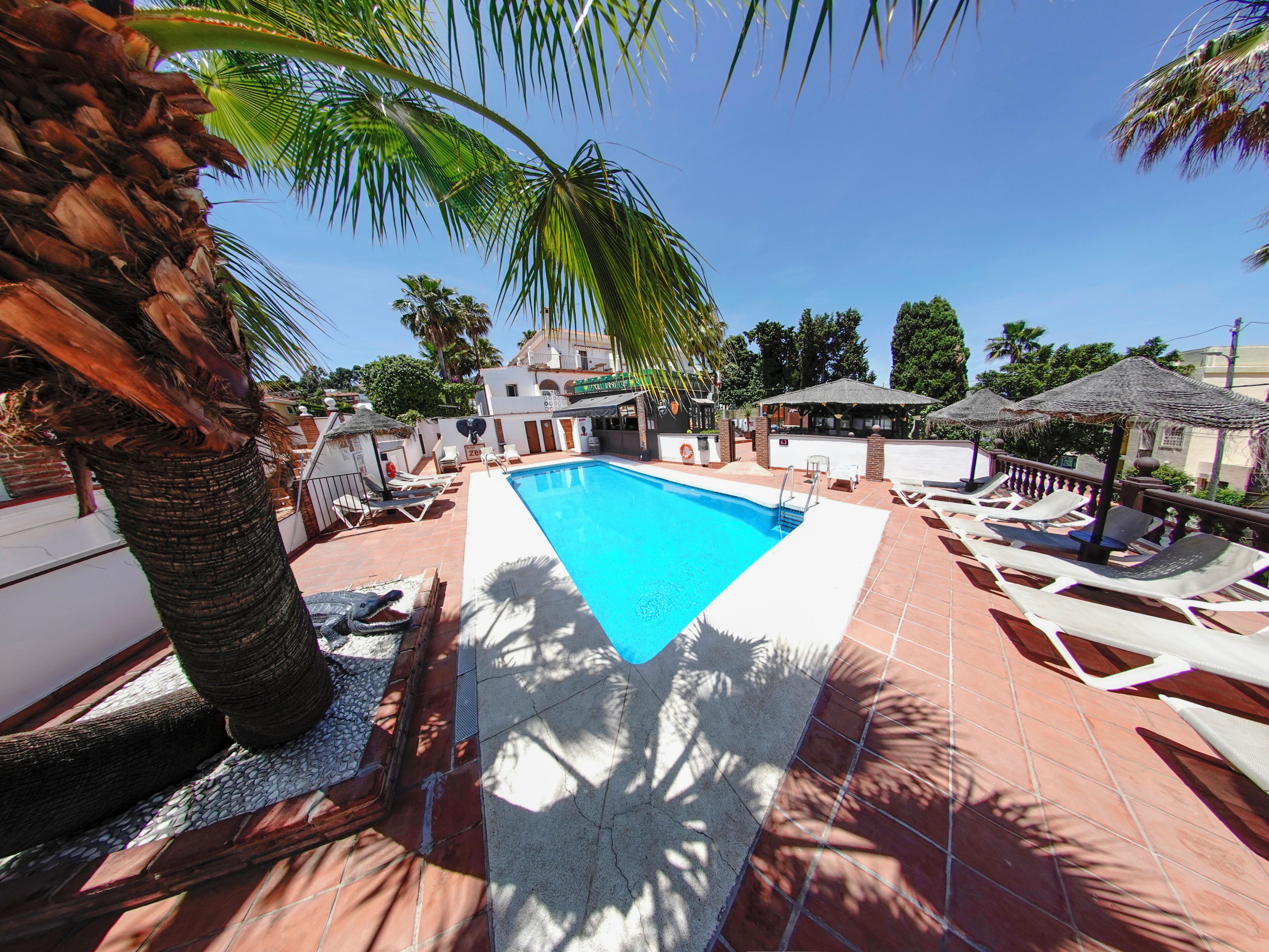 A swimming pool with lounge chairs and umbrellas, surrounded by palm trees and a building with a tiled roof.