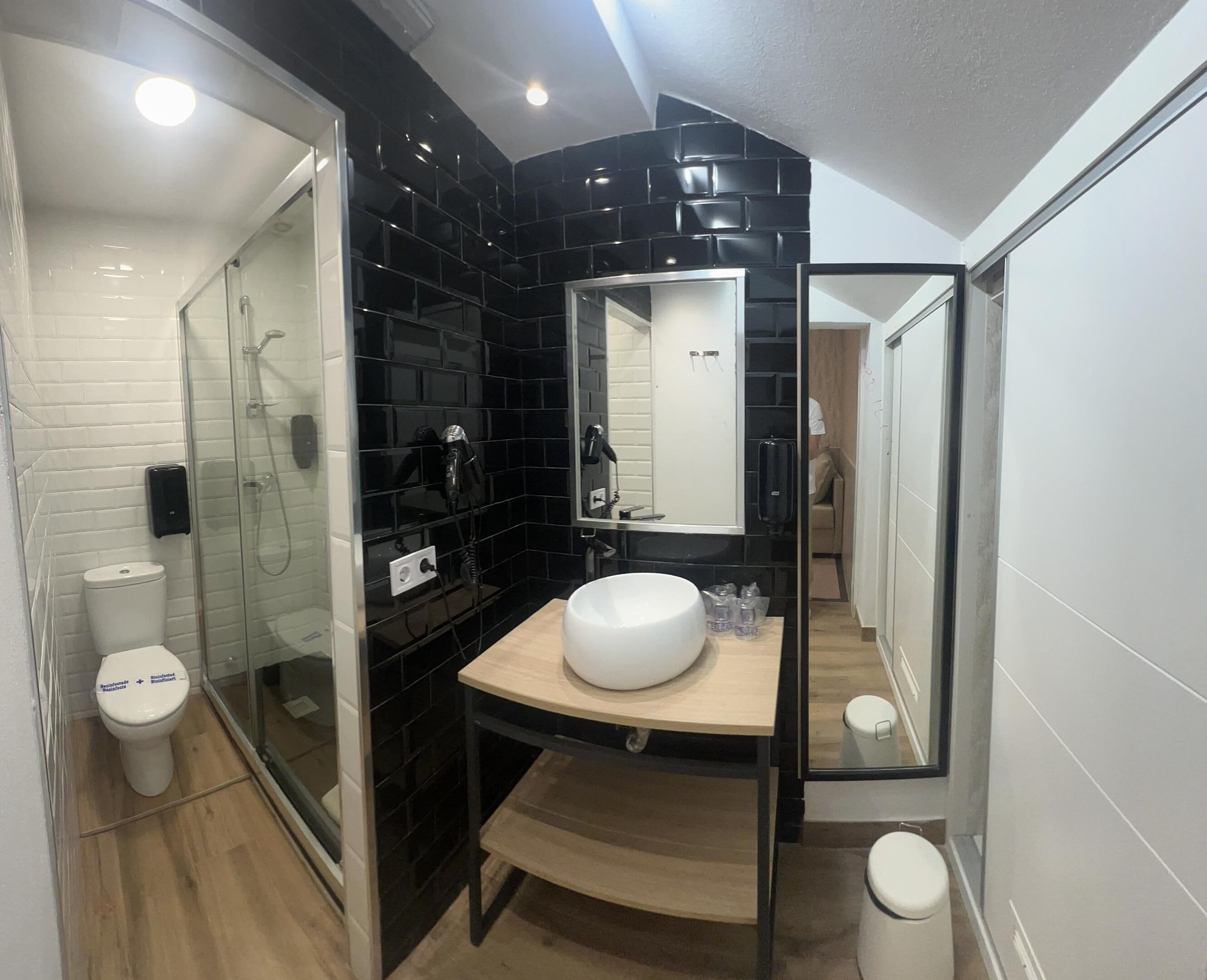 Un baño moderno con cabina de ducha de vidrio, inodoro blanco, pared de azulejos negros y tocador de madera con lavabo blanco.