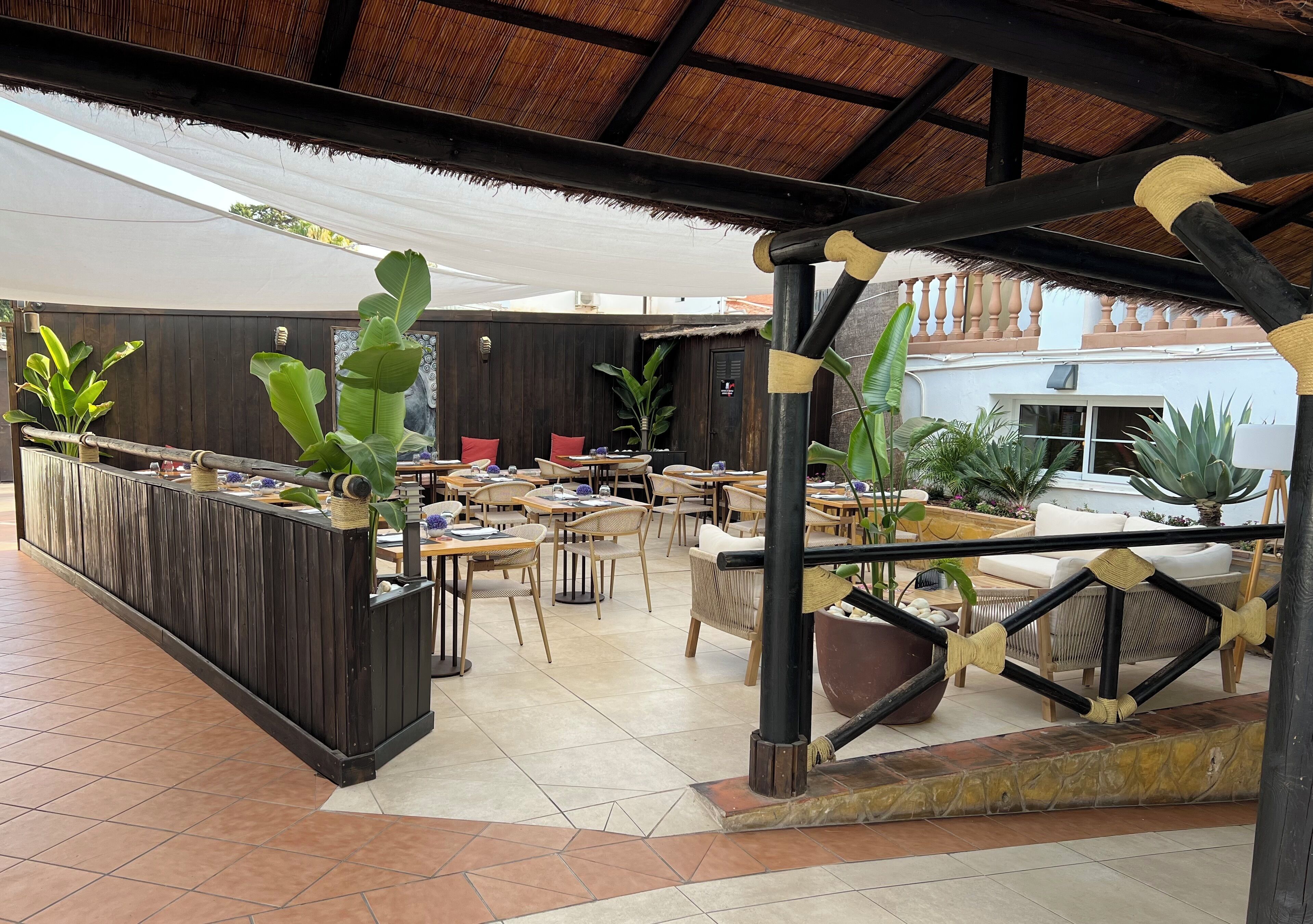 Un área de comedor al aire libre con mesas y sillas de madera, rodeada de plantas en macetas y una estructura de techo de paja.