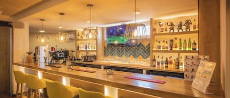 A bar area with wooden bar counter, yellow bar stools, hanging pendant lights, and shelves with bottles.