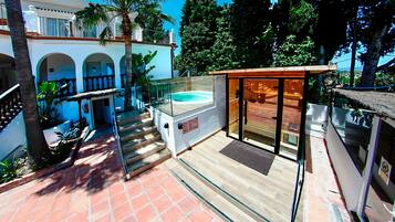 Un área de piscina con cerramiento de vidrio, terraza de madera y escalera que conduce a un espacio de estar cubierto al aire libre.