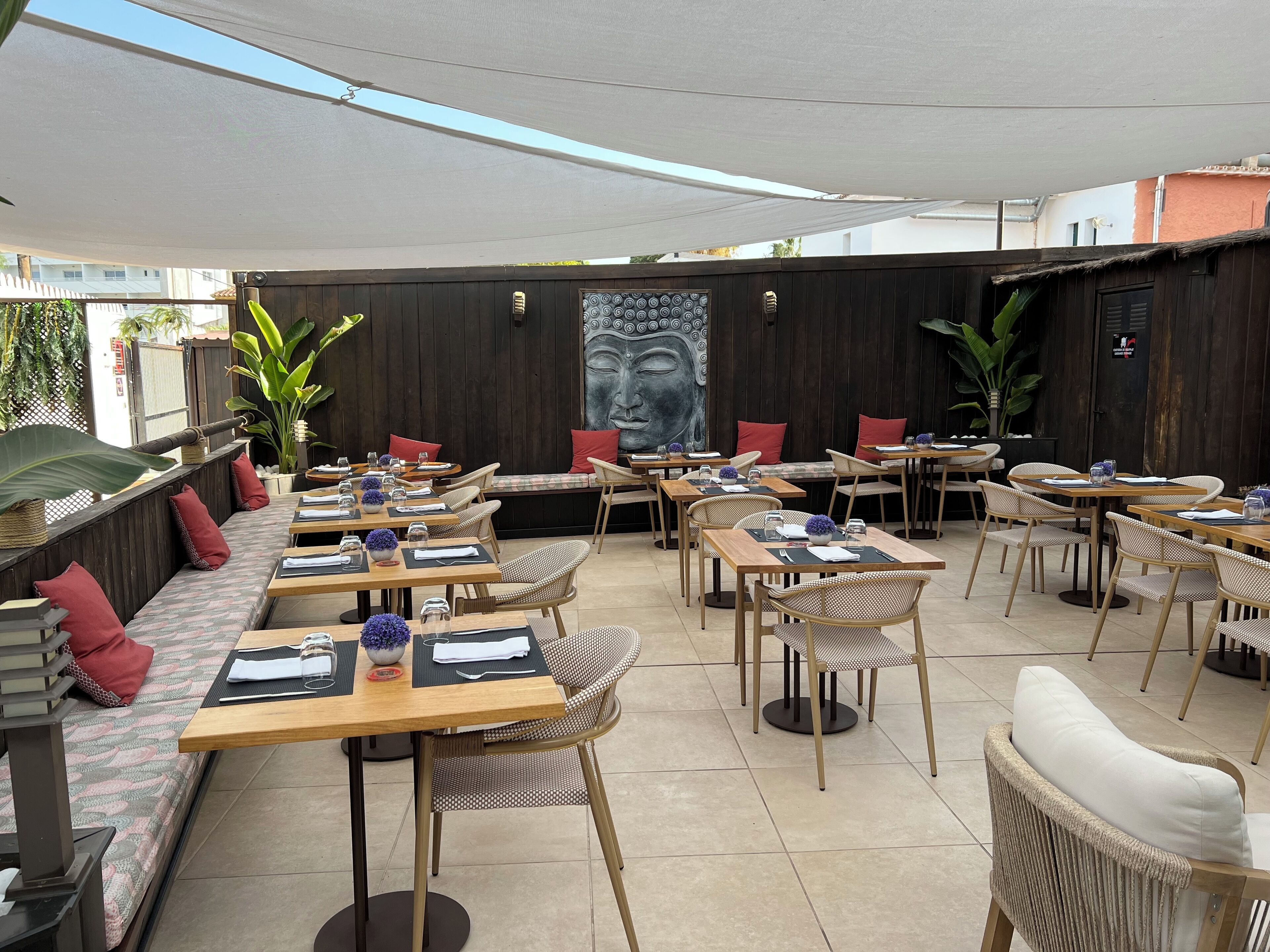 An outdoor dining area with wooden tables and wicker chairs, some with cushions. There are potted plants and a Buddha mural on the wall.