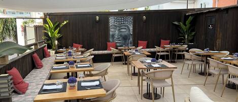 An outdoor dining area with wooden tables and wicker chairs, some with cushions. There are potted plants and a Buddha mural on the wall.