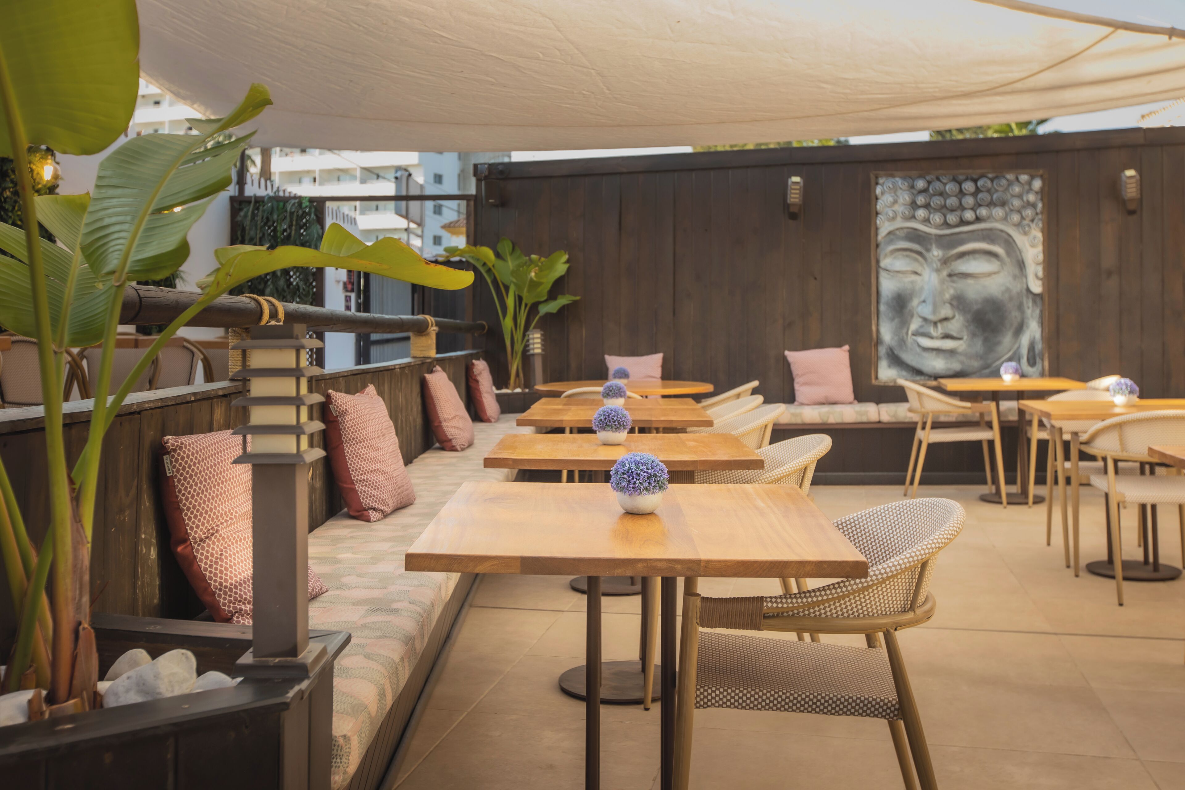 An outdoor seating area with wooden tables and wicker chairs, a Buddha mural on the wall, and large potted plants.