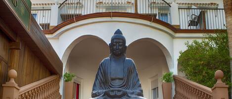 Une statue de Bouddha se trouve dans une cour dont le sol est recouvert de carreaux, entourée d’un escalier et d’un bâtiment doté de balcons.