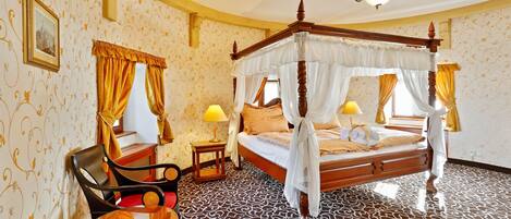A four-poster canopy bed with a wooden frame and white drapes, a wooden chair with a cushion, a small bedside table with a lamp, and patterned wallpaper.
