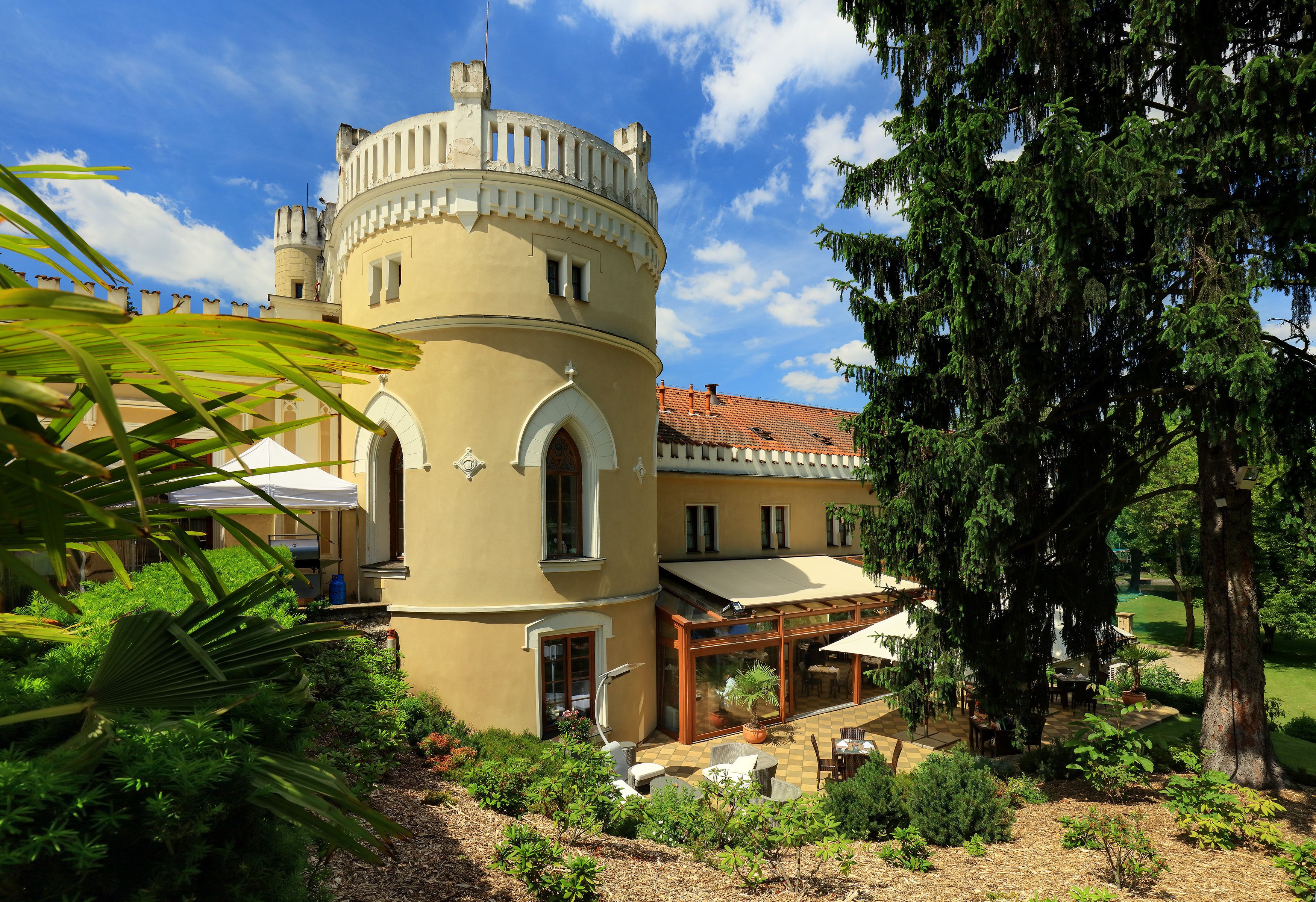 Un edificio de estilo castillo con una torre redonda, un patio con mobiliario exterior y un jardín con diversas plantas.