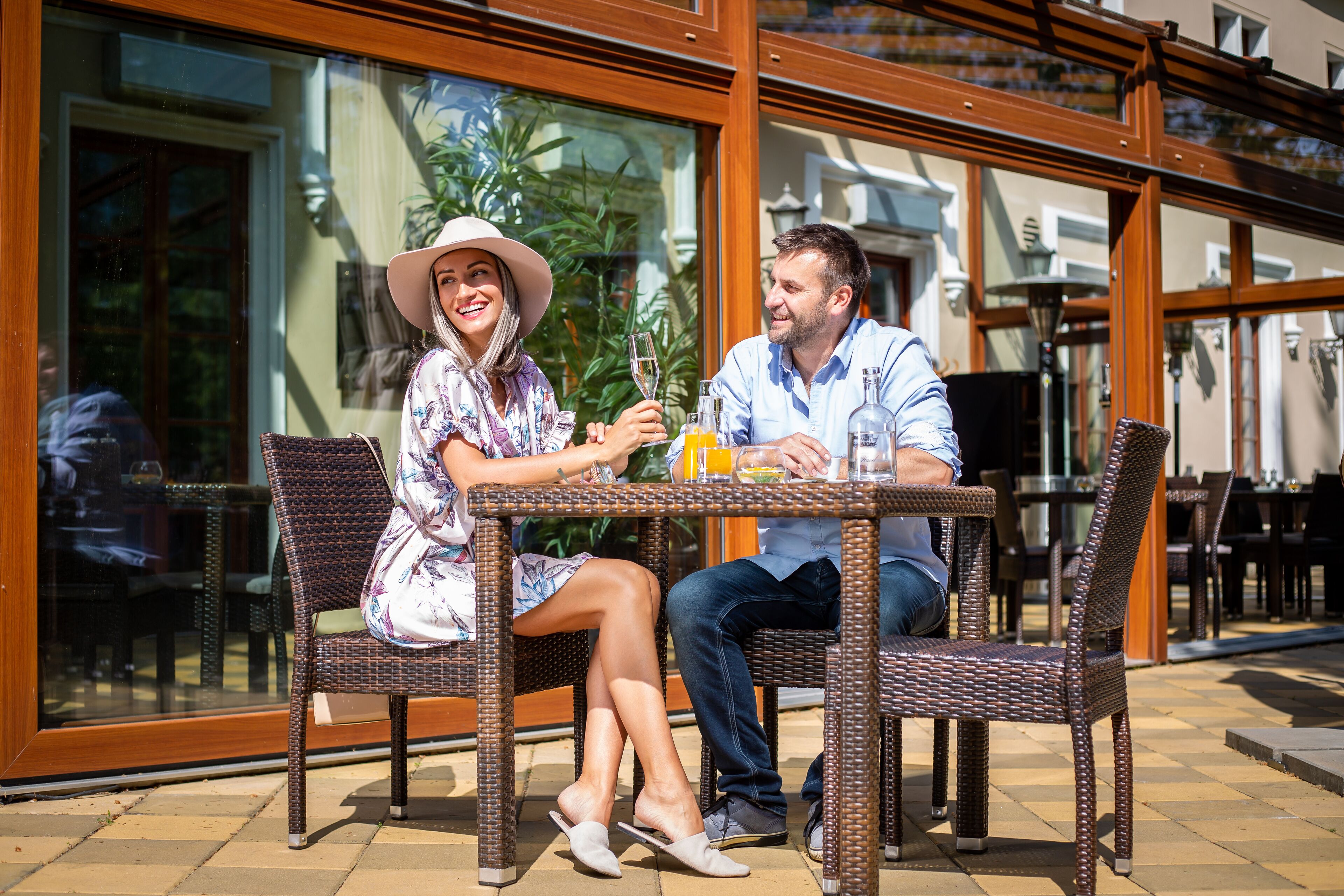 A couple seated at an outdoor table with wicker chairs, a glass window in the background, and a tiled floor.