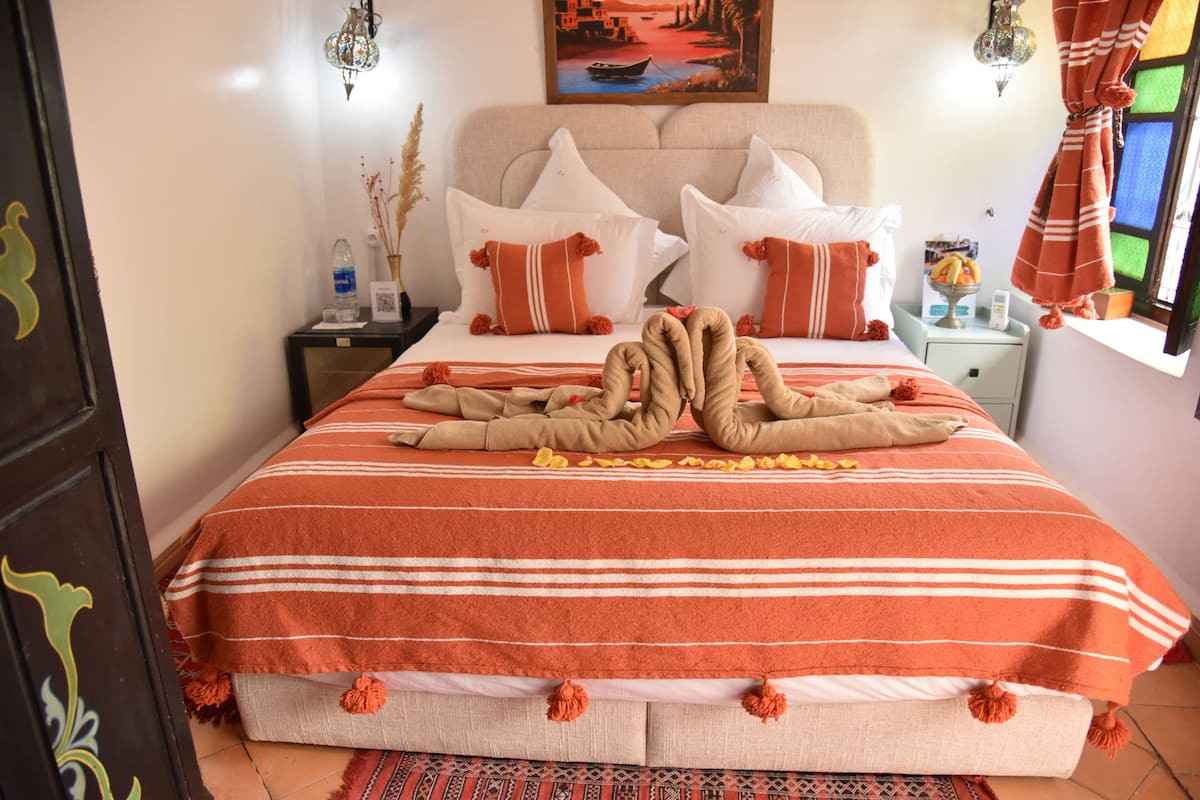 Un lit bien fait, avec une couverture à rayures et des coussins décoratifs, une table de chevet sur laquelle se trouvent une bouteille et un cadre photo, et une fenêtre avec des rideaux.