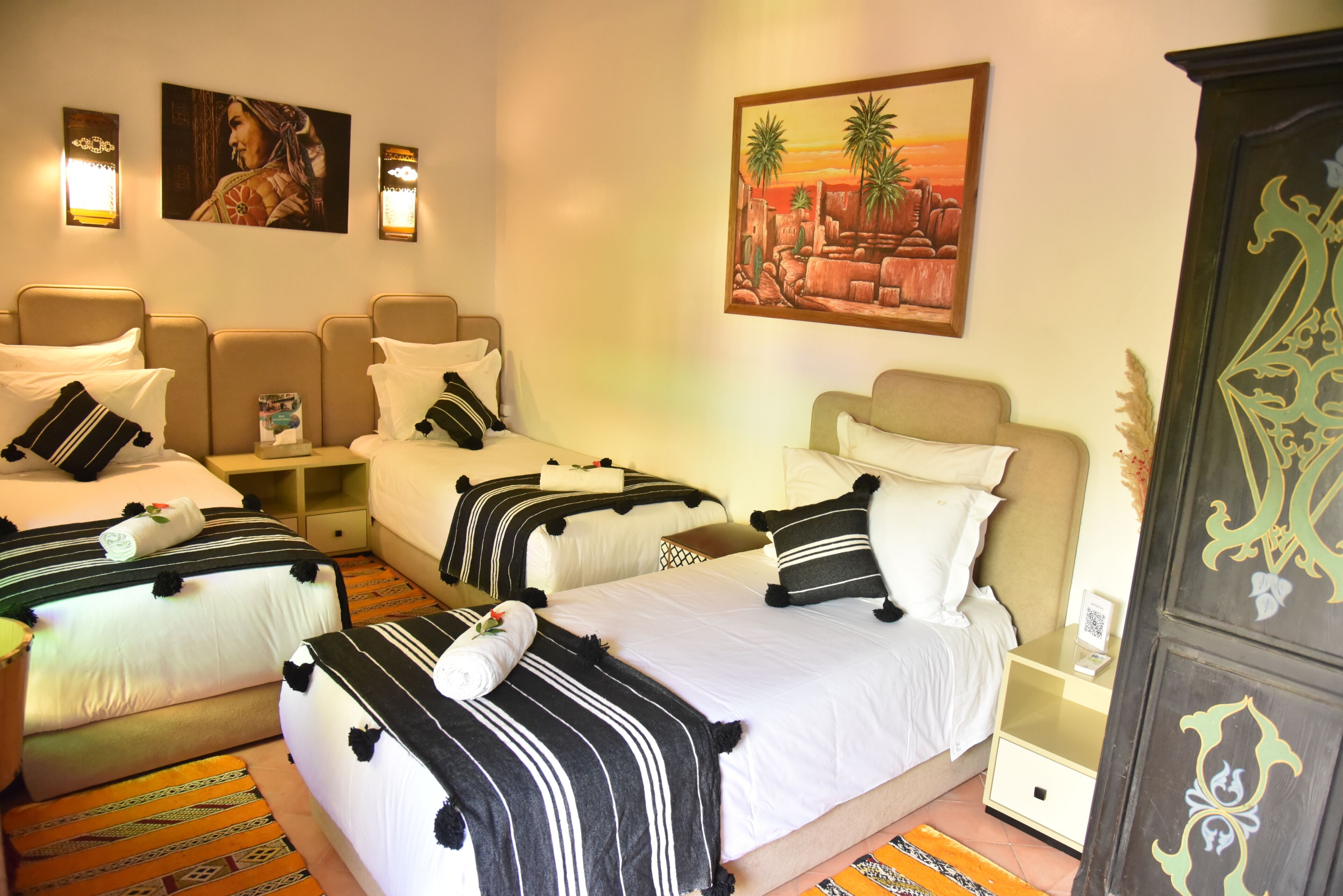 A hotel room with three beds, each with black and white striped bedding and matching pillows. There are wall-mounted lights and a painting of a desert landscape.