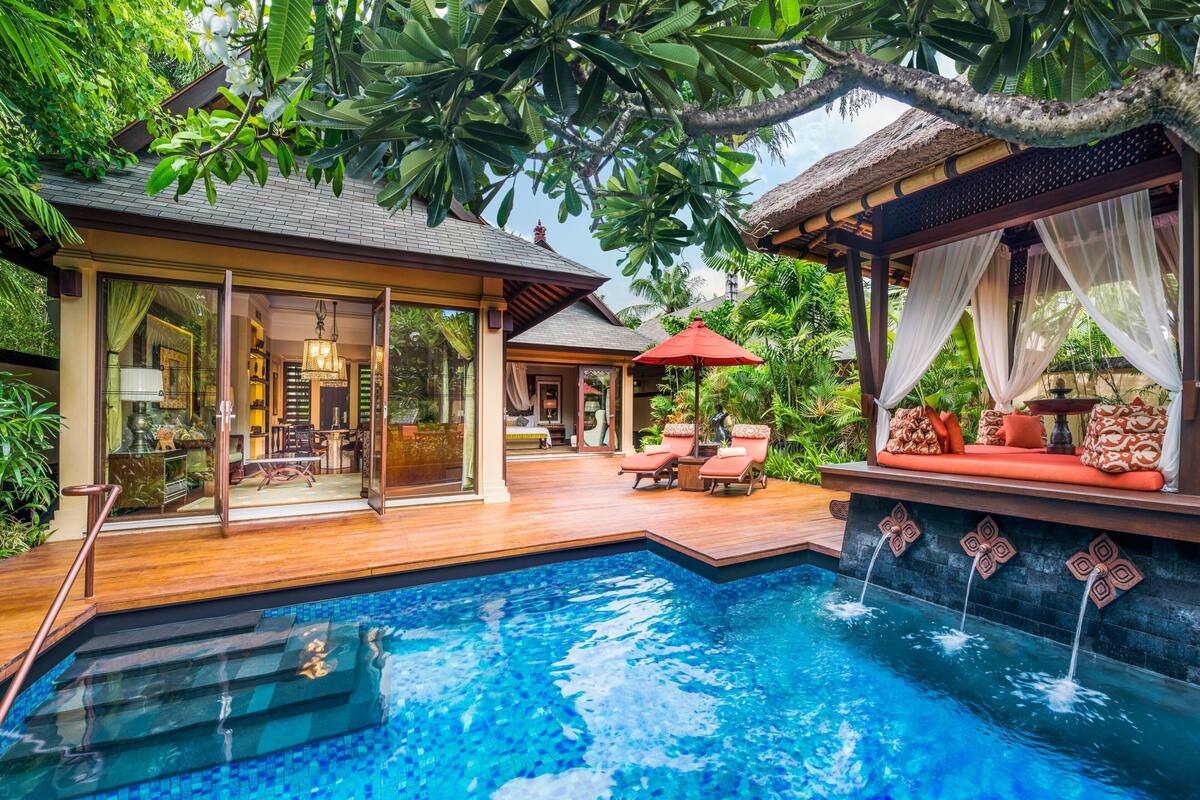 Un espace piscine avec une terrasse en bois, un coin salon doté de coussins rouges et un espace détente couvert avec un parasol rouge.