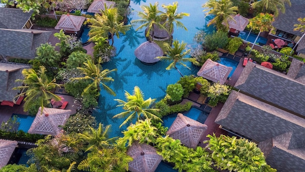 Ett flygbildsfoto av ett semesterresort med flera bungalows som är byggda på pålar över vattnet, en central pool och en solnedgång över havet.