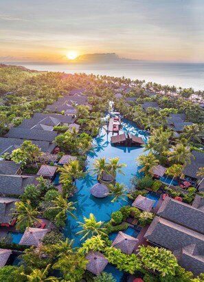 An aerial view of a resort with multiple overwater bungalows, a central pool, and a sunset over the ocean. - The St. Regis Bali Resort (Nusa Dua)