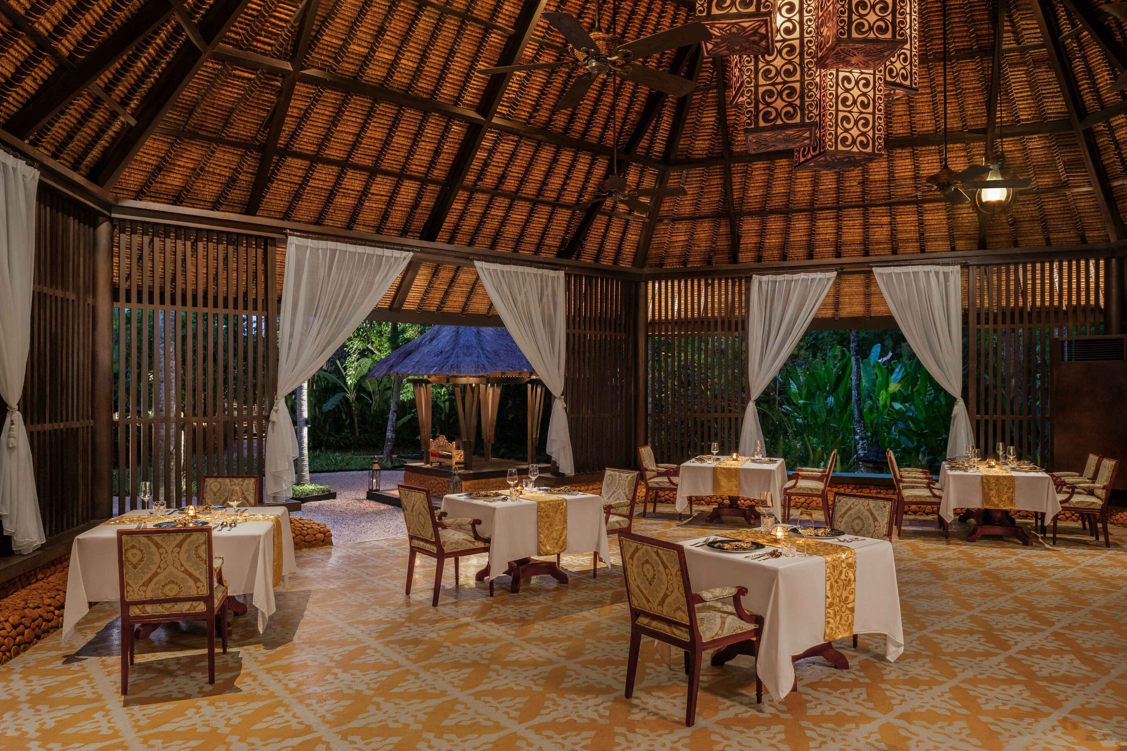 An indoor dining area with tables set for a meal, featuring a thatched roof and natural light from large windows.