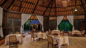 An indoor dining area with tables set for a meal, featuring a thatched roof and natural light from large windows.