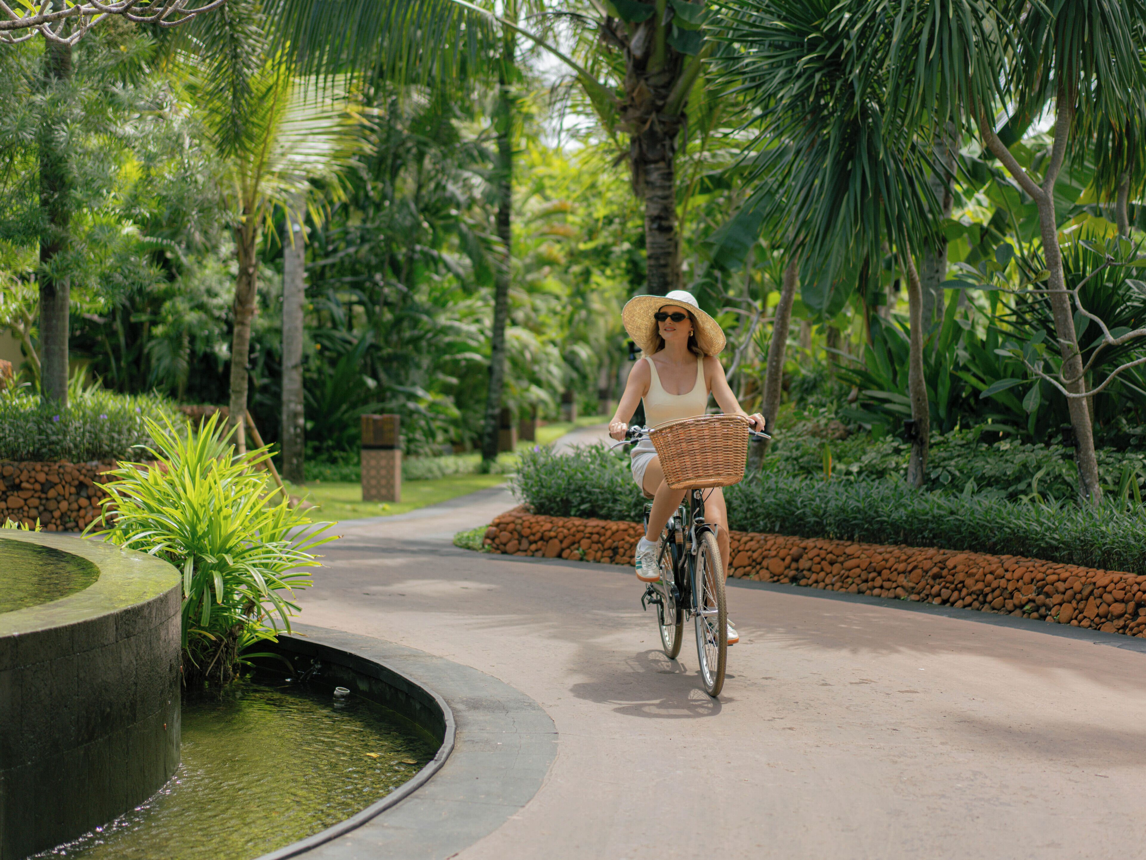 Una mujer pedaleando en bicicleta por un camino pavimentado, rodeada de exuberante vegetación y palmeras.