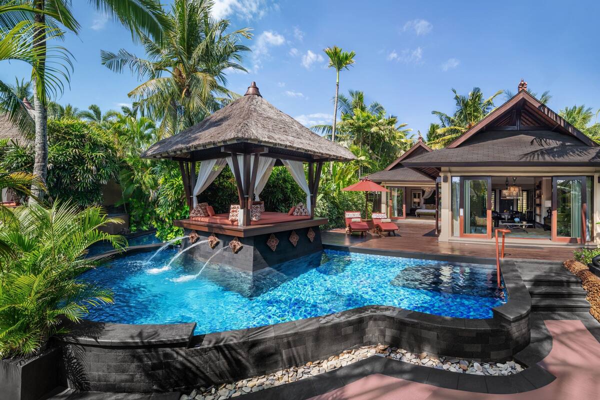 Un espace piscine agrémenté d’un gazebo à toit de chaume, de chaises longues et d’un bâtiment doté de grandes fenêtres.