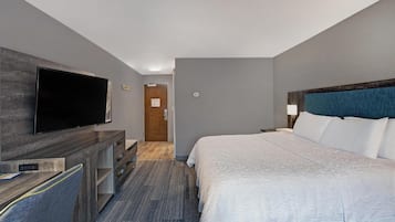 A hotel room with a bed, a TV mounted on a wooden dresser, a desk with a chair, and a door leading to another room.