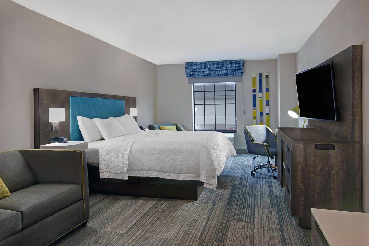A hotel room with a bed, a desk, a chair, a sofa, a TV, and a window.