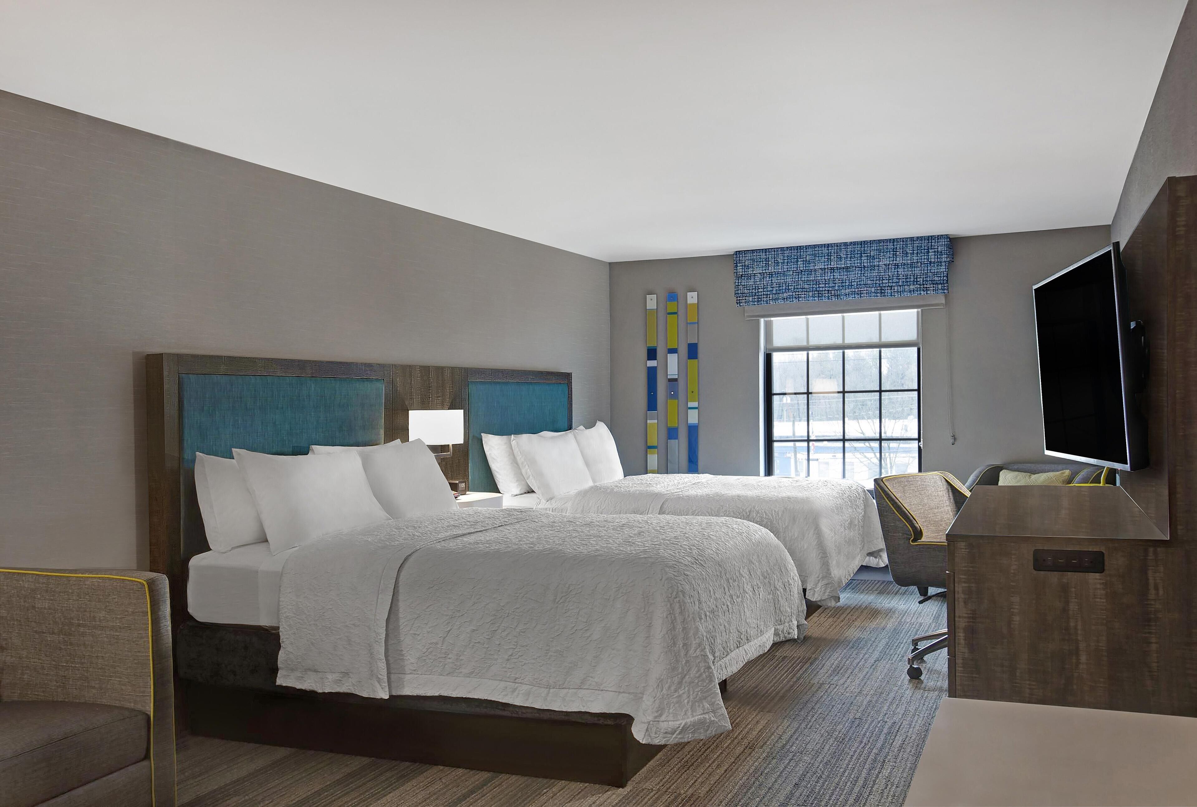 A hotel room with two beds, a desk, a TV, and a window with blinds.